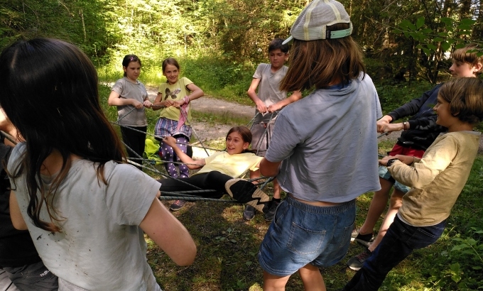 Kinder auf einem Waldweg, in der Mitte liegt ein Kind auf einem aufgespanntem Netz