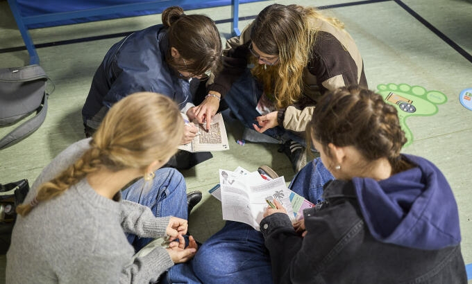 Vier Jugendliche sitzen auf dem Boden im Kreis und schauen auf Papiere.